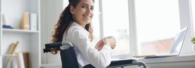 Woman in wheelchair smiling while holding a coffee cup at her desk.